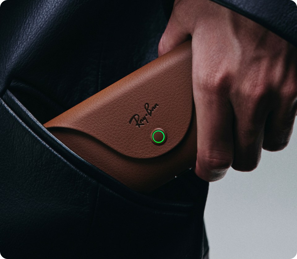 A hand holding a brown Ray-Ban charging case with a green status light.